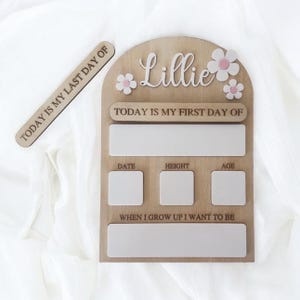 May include: Wooden milestone board with the name "Lillie" and flower accents. Includes sections for "Today is my first day of," date, height, age, and "When I grow up I want to be." A separate wooden piece reads "Today is my last day of."