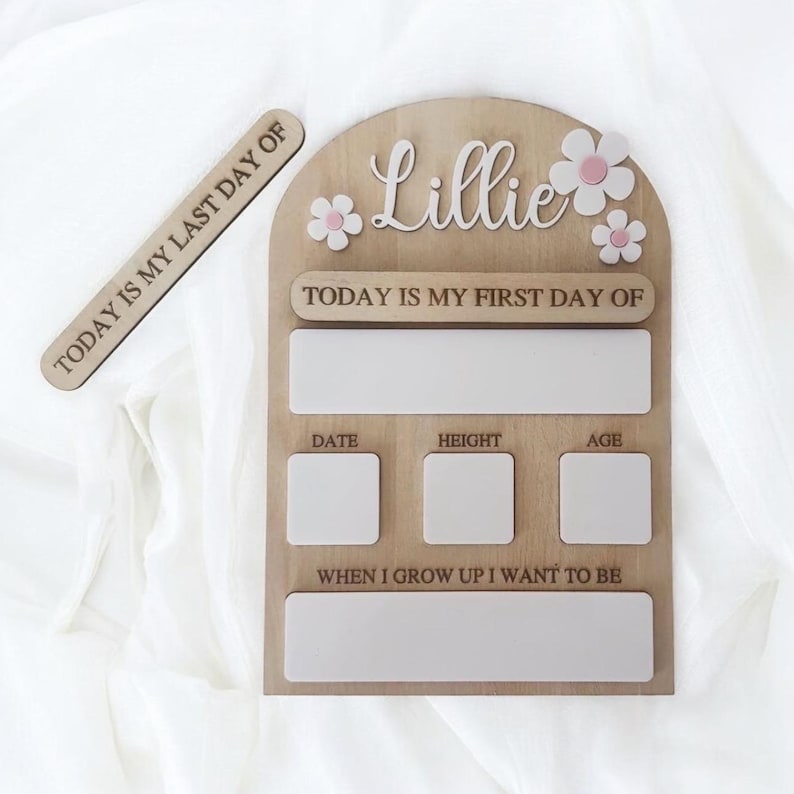 May include: Wooden milestone board with the name "Lillie" and flower accents. Includes sections for "Today is my first day of," date, height, age, and "When I grow up I want to be." A separate wooden piece reads "Today is my last day of."