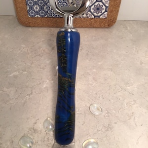 May include: A bottle opener with a shiny silver top and a blue handle with a gold and green design. The bottle opener is standing on a clear plastic stand. Decorative glass pebbles are scattered around the base.