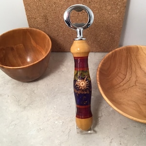 May include: A wooden bottle opener with a chrome bottle opener on top. The handle is decorated with a colourful floral design in shades of blue, green, red, and yellow.