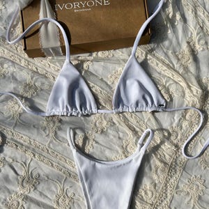 May include: White bikini top and bottom with thin straps. The top is a triangle style and the bottom is a high-cut style. The bikini is on a white lace fabric background.