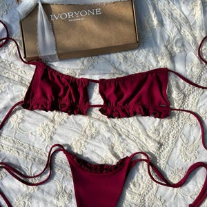 Puede incluir: Un top y una parte inferior de bikini burdeos con ribete de volantes. El top tiene un detalle de corte en la parte delantera y se ata en el cuello y la espalda. La parte inferior es de estilo de corte alto con lazos a los lados.