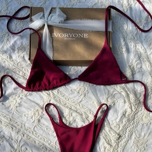 Peut inclure: Un ensemble de bikini bordeaux avec un haut triangle et un bas taille haute. Le bikini est emballé dans une boîte avec le nom de la marque "IVORYONE SWIMWEAR" imprimé dessus.