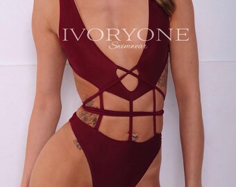 May include: A deep burgundy one-piece swimsuit featuring a plunging V-neckline, cut-out sides, and a strappy midsection design. The text IVORYONE Swimwear is printed in white across the chest area of this solid-colored, high-cut swimwear garment.