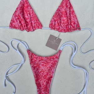 Könnte beinhalten: Zweiteiliger Bikini mit floralem Muster in Rosa- und Weißtönen. Das Oberteil ist im Triangel-Stil mit weißen Trägern gehalten, das Unterteil im String-Stil. Das Etikett lautet "IVORYONE".