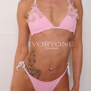 May include: A light pink bikini with floral embellishments. The triangle top has white straps and is adorned with delicate, light-colored flowers. The matching bottoms have tie sides. The brand name "IVORYONE Swimwear" is visible.