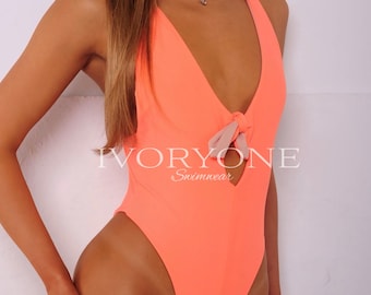 Op de afbeelding: Een neonoranje badpak uit één stuk met een diepe V-hals, een centraal knoopdetail en een kleine uitsnijding. Het kledingstuk toont de tekst IVORYONE Swimwear in witte letters op de borst en heeft een hoog uitgesneden beenontwerp.