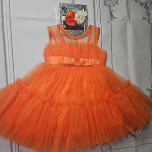 May include: A bright orange tulle dress with a large bow at the waist. The dress has multiple layers of tulle and is perfect for a special occasion.
