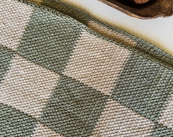 Könnte beinhalten: Eine gehäkelte Decke in Grün und Beige mit einem karierten Muster. Die Decke ist in der Hälfte gefaltet und liegt auf einer weißen Oberfläche. In der oberen linken Ecke des Bildes befinden sich Wollknäuel in einer Holzschale.