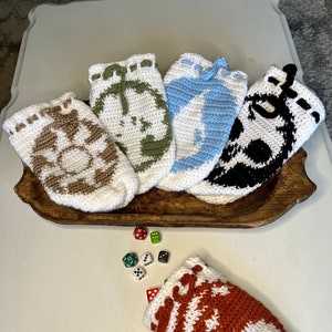 Op de afbeelding: Vijf gehaakte dobbelstenen zakken met verschillende ontwerpen, waaronder een schedel, een halve maan, een anker, een bloem en een pompoen. De zakken liggen op een houten dienblad met dobbelstenen eromheen.