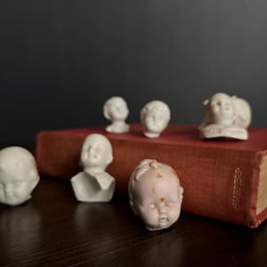 May include: A collection of antique doll heads in various sizes and styles, arranged on and around a vintage red book. The doll heads are primarily white porcelain, with some showing signs of age and wear.