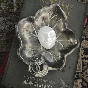 May include: A silver-toned leaf-shaped dish holds a small white face sculpture. The dish rests on a book with the title "MONSTERS" and the author's name, "ADAM BENEDICT". The background has a patterned, textured appearance.