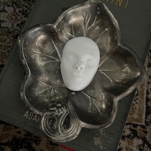 May include: A silver-toned, leaf-shaped dish holding a small, white, sculpted face. The dish sits atop a dark green book with gold lettering that reads "MONSTERS IN PRINT". The dish has intricate detailing.
