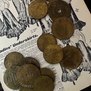 May include: A collection of vintage brass tokens with various designs and text, including "No. 31R387 69c", "Hungry Eye Saloon", "Good For $2.00", and "Just Dooie".