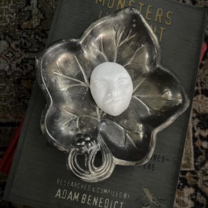 May include: A silver leaf-shaped dish holds a small white face sculpture. The dish rests on a dark green book titled "Monsters" with the author's name, Adam Benedict, visible.