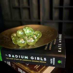 May include: A brass-colored decorative bowl filled with translucent green glass marbles. The bowl sits atop a stack of books, including "The Radium Girls" by Kate Moore. The books are on a dark surface.