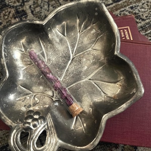 May include: A silver-toned metal dish shaped like a leaf with embossed veins and a cluster of grapes at the base. Resting inside the dish is a clear glass vial sealed with a cork stopper, containing small, dried purple floral buds or petals.