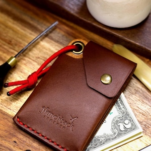 May include: A brown leather wallet with a red stitching detail and a red cord. The wallet has a flap with a brass button closure. The wallet has the text "Hunchback Leather" embossed on the front. The wallet is sitting on a wooden surface with a few US dollar bills inside.