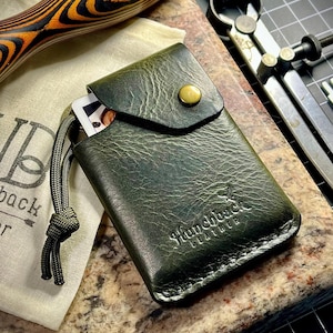 May include: A green leather card holder with a flap and a button closure. The holder has a stitched edge and the text "Hunchback Leather" embossed on the front.