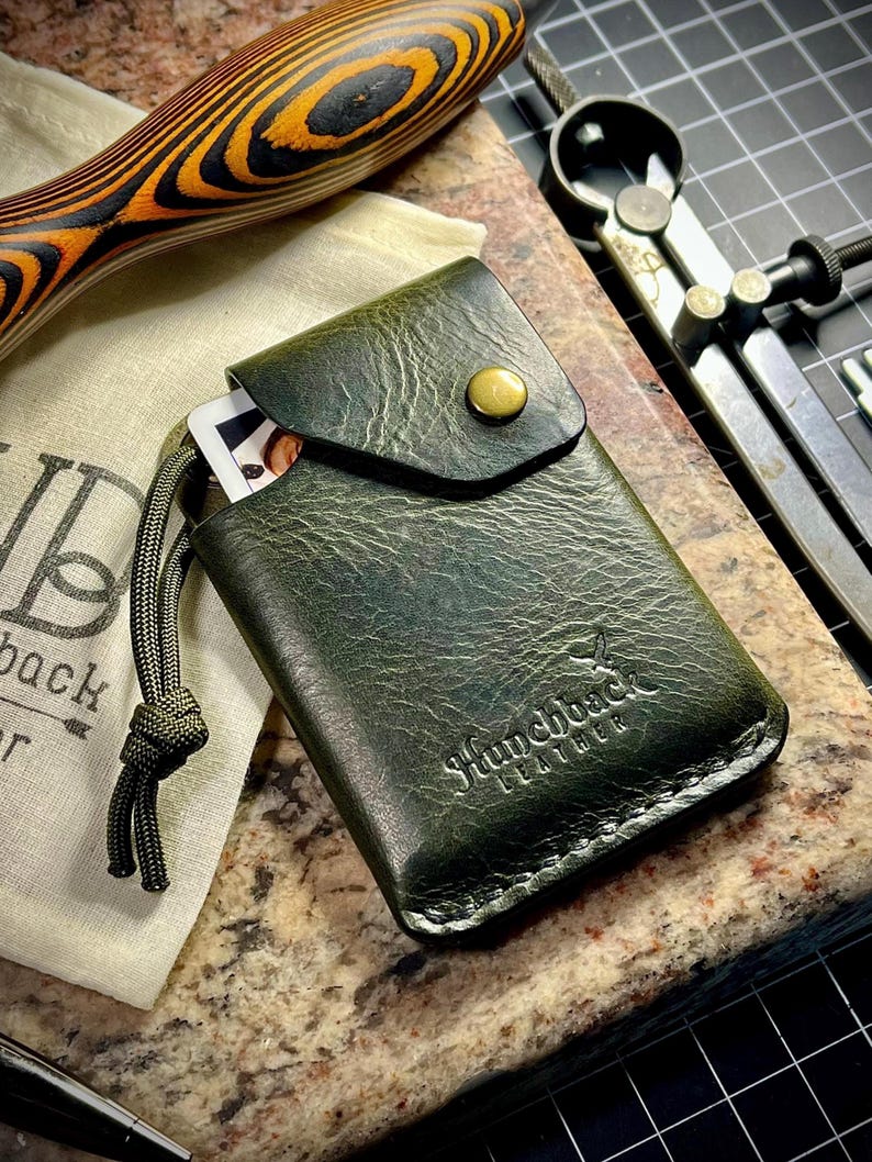 May include: A green leather card holder with a flap and a button closure. The holder has a stitched edge and the text "Hunchback Leather" embossed on the front.