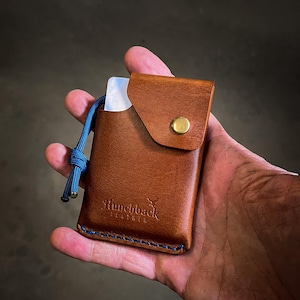 May include: A brown leather card holder with a blue cord and a brass button. The card holder has a flap with a stitched edge and the text "Hunchback Leather" embossed on the front.