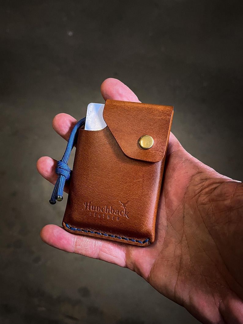 May include: A brown leather card holder with a blue cord and a brass button. The card holder has a flap with a stitched edge and the text "Hunchback Leather" embossed on the front.