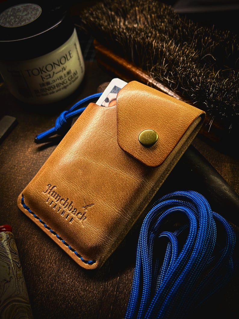 May include: A tan leather card holder with a blue cord and a brass button. The card holder has the text "Hunchback Leather" embossed on the front.