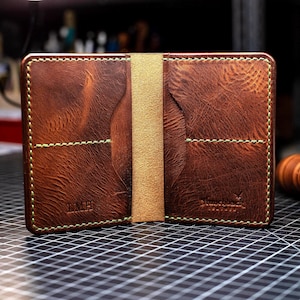 May include: A brown leather bifold wallet featuring a textured surface, light green contrast stitching, and two interior card slots on each side. The left side is embossed with the letters LMH, and the right side features a small logo stamp.
