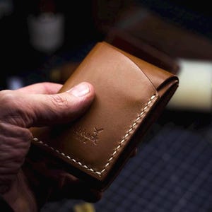May include: A brown leather wallet with white stitching and a logo. The wallet is held in a hand, showcasing its compact design. The stitching is visible along the edges, and the logo is embossed on the front.