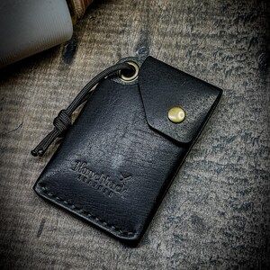 May include: Black leather wallet with a fold-over flap and a brass button closure. The wallet has a stitched edge and the text "Hunchback Leather" embossed on the front.