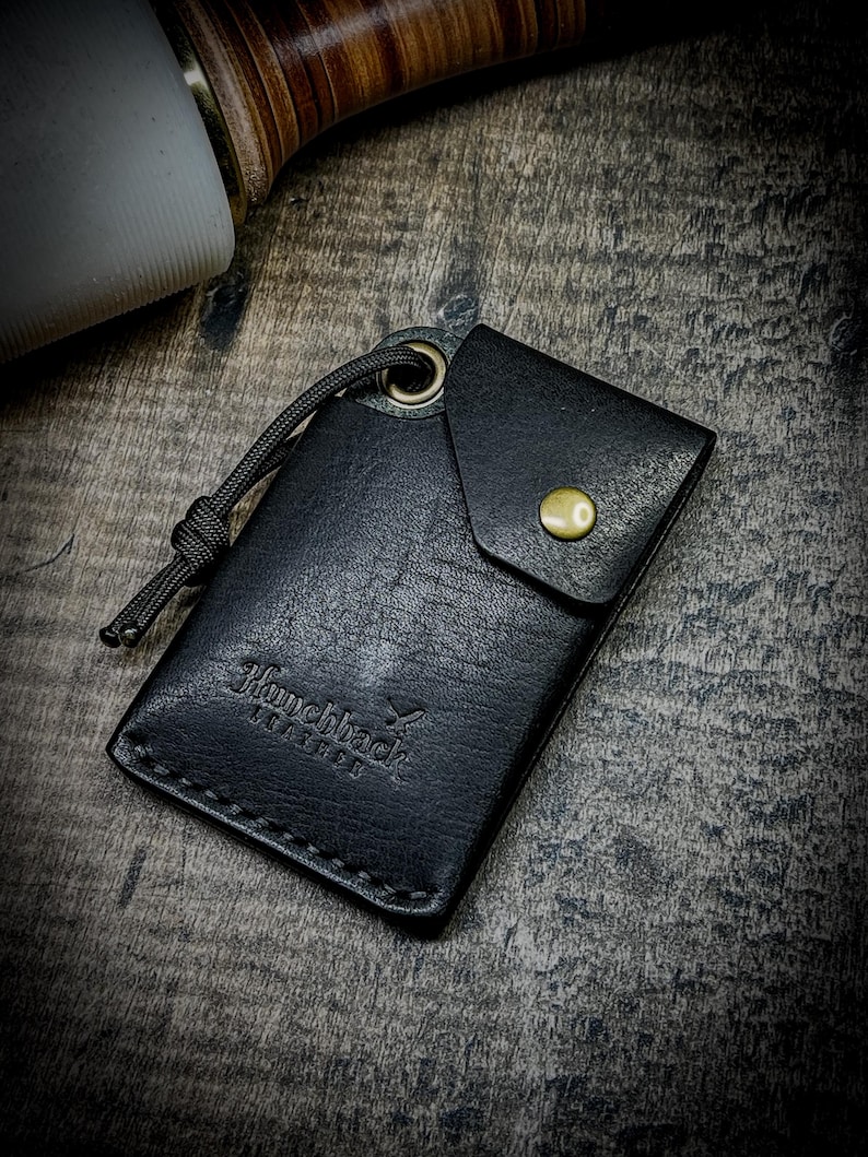 May include: Black leather wallet with a fold-over flap and a brass button closure. The wallet has a stitched edge and the text "Hunchback Leather" embossed on the front.