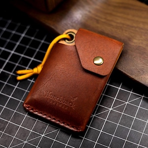 May include: A brown leather key chain with a small pocket and a yellow cord. The key chain has the text "Sharpback Leather" embossed on it.