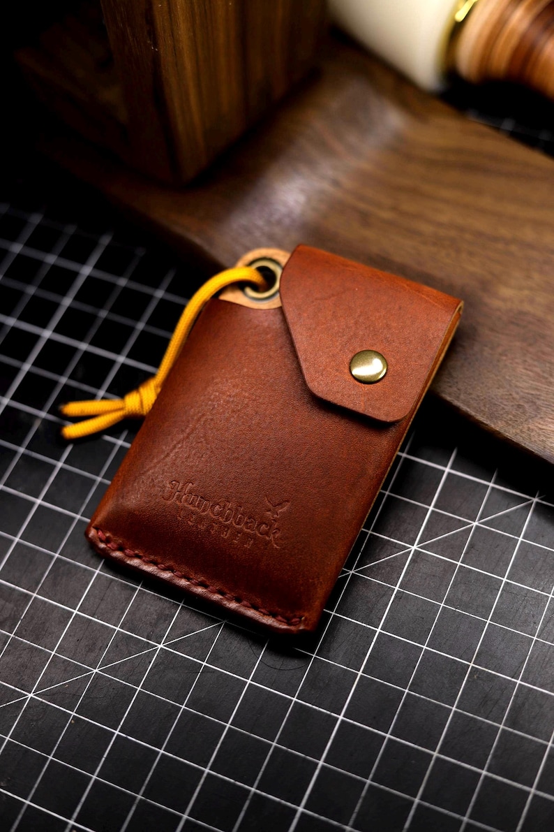 May include: A brown leather key chain with a small pocket and a yellow cord. The key chain has the text "Sharpback Leather" embossed on it.