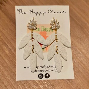 Könnte beinhalten: Weiße Keramikohrringe mit einem goldenen Blattdesign und einem Kristallkronendetail. Die Ohrringe werden auf einer weißen Karte mit dem Text "The Happy Clover" und einer Website-Adresse präsentiert.