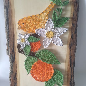 May include: String art of a white flower with yellow center, two orange fruits with green leaves, and a yellow bird on a wooden background.