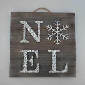 Puede incluir: Un letrero de madera cuadrado con la palabra "NOEL" en arte de hilo blanco y un copo de nieve blanco. El letrero tiene un acabado de vetas de madera gris-marrón y una cuerda de yute para colgar. Decoración navideña.