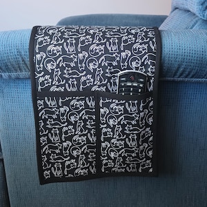 May include: Black and white patterned armrest organizer with a cat design. The organizer has two pockets and a pocket holding a remote control. The organizer is placed on a blue recliner.
