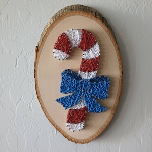 May include: A string art piece on an oval wood slice featuring a candy cane design created with red and white thread wrapped around silver nails. A blue thread bow is attached to the center of the candy cane, adding a contrasting color element.