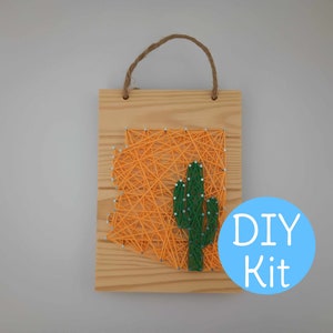 May include: DIY string art kit featuring the outline of Arizona with a green cactus in the centre. The kit is on a wooden base with a rope hanger.
