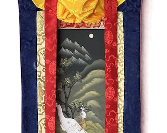 May include: A traditional Tibetan thangka painting depicting a white elephant with a monkey and rabbit riding on its back. The painting is framed with a blue, red, and gold border and has a red and gold patterned fabric at the bottom.