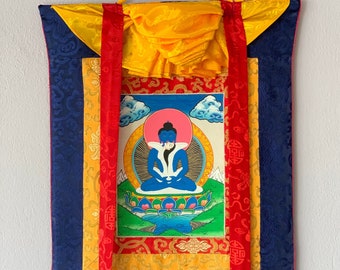 May include: A vibrant, ornate wall hanging featuring a central image of a seated blue figure with a pink halo. The artwork is framed by a yellow and red border, with a deep blue patterned fabric background. Red ribbons hang from the top.