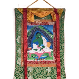 May include: A traditional Tibetan thangka painting on silk depicting a green deity seated in a cave. The thangka is framed with a decorative border and has red silk ties for hanging.