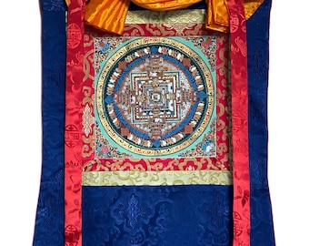 May include: A Tibetan prayer flag featuring a colourful mandala design. The flag is made of silk and has a red, gold and blue border. It is hung on a wooden dowel.