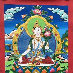 Pode incluir: Uma pintura colorida de uma divindade sentada em uma postura meditativa. A divindade está cercada de flores e tem uma auréola dourada. A pintura está sobre um fundo vermelho.