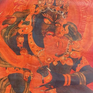 May include: A vibrant red and dark blue thangka painting depicting a wrathful deity in a fierce embrace with a consort. The figures are adorned with intricate jewelry, crowns, and skulls, set against a fiery red background with detailed line work.