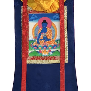 May include: A traditional Buddhist thangka painting depicting a blue Buddha figure in a meditative pose. The artwork features vibrant colors, including gold, red, blue, and orange, and is framed with ornate fabric and wooden dowels. The painting is approximately 30 inches in height.