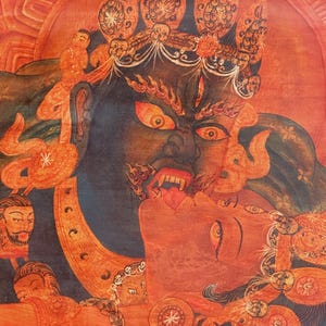 May include: A detailed thangka painting featuring a dark blue deity with a fierce expression, wide orange eyes, and bared teeth, embracing a red-skinned figure. The composition includes ornate golden crowns, skull motifs, and smaller faces.