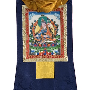 Può includere: Un tradizionale dipinto thangka tibetano, con una figura centrale dai colori vivaci. L'opera d'arte è incorniciata con bordi ornati e posta su uno sfondo di broccato blu scuro. La pergamena include tasselli di legno e una parte superiore in tessuto dorato.