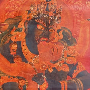May include: A vibrant thangka painting featuring a dark blue deity with a fierce expression and bared teeth, embracing a red figure. The composition includes intricate golden crowns, jewelry, and a circular halo against a deep red background.