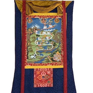 Può includere: Un arazzo thangka verticale con un dipinto centrale raffigurante un sentiero tortuoso con figure e animali, incorniciato da bordi in tessuto rosso e oro. L'opera è montata su uno sfondo in broccato di seta blu scuro con un cordino dorato.